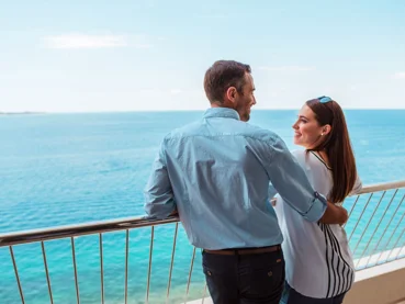 Moški in ženska se nasmejano objemata ob ograji terase s pogledom na morje