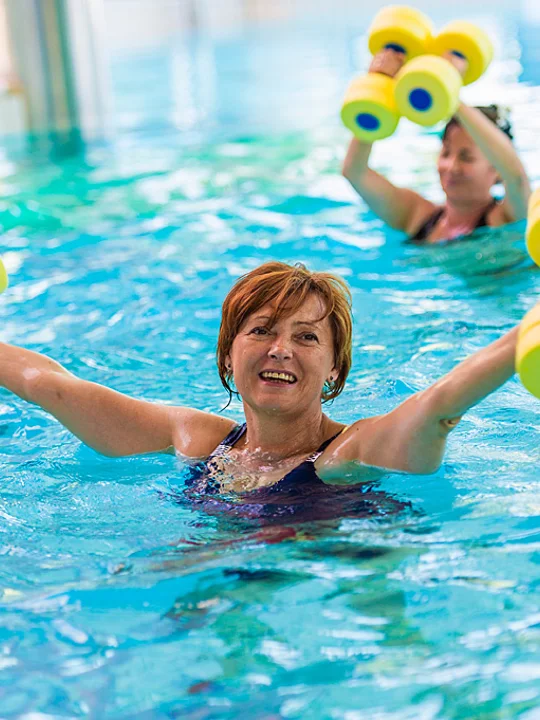 Ženska v srednjih letih se v notranjem bazenu rekreira z rumenimi utežmi v roki