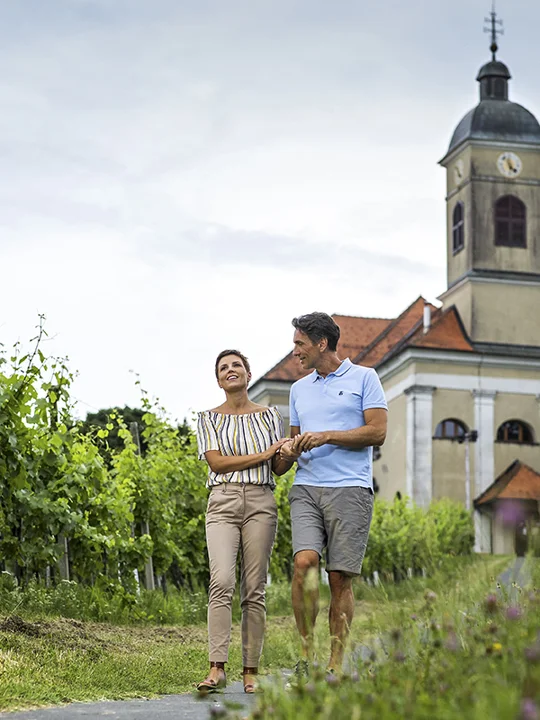 Par v srednjih letih hodi po potki iz cerkve, na levi in desni strani so zeleni vinogradi
