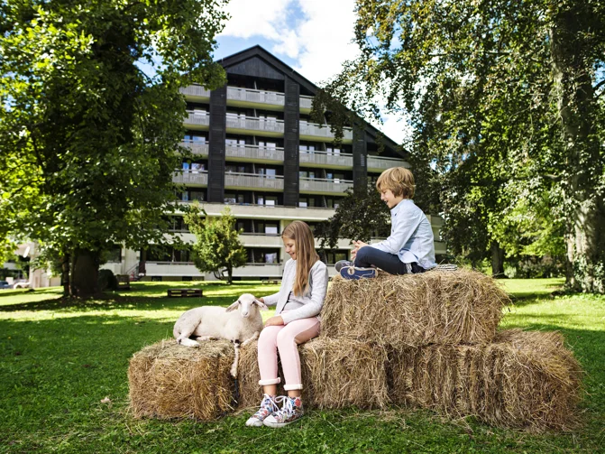 Deček in deklica sedita na kupu sena pred hotelom na travnati površini in božata ovco