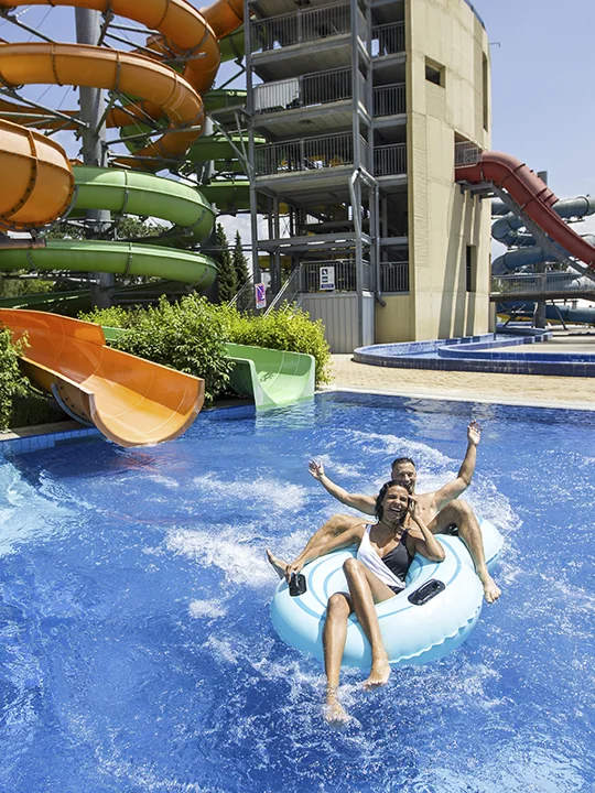 Mlad par se na modri blazini spušča iz oranžnega tobogana v zunanji bazen