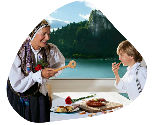 Ženska v narodni noši s kuhalnico v roki pogleduje proti otroku, ki ima pred seboj na pladnju mesne dobrote in v rokah kos klobase