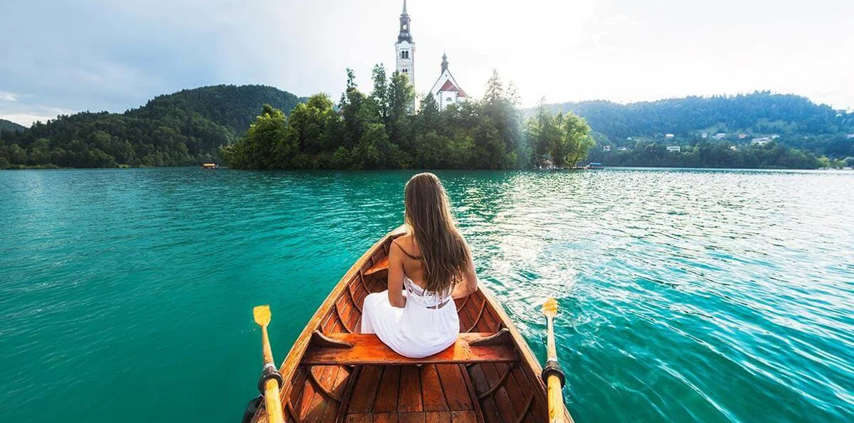 Sava Hotels & Resorts Ženska v beli obleki sedi na čolnu na blejskem jezeru in gleda cerkev na otoku