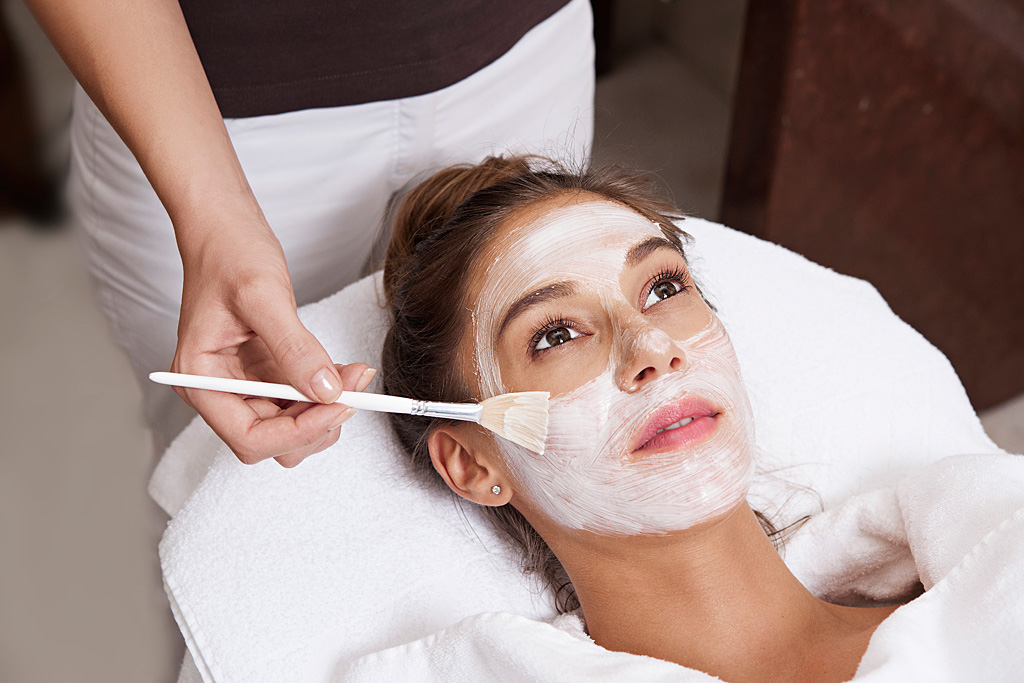 Ženska, ki leži na masažni mizi in ji kozmetičarka nanaša zdravilno obrazno masko bele barve