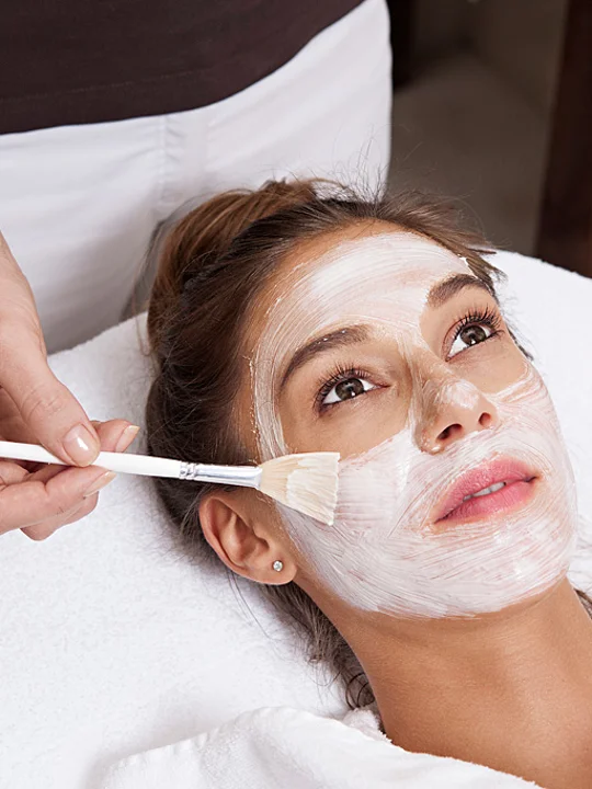 Ženska, ki leži na masažni mizi in ji kozmetičarka nanaša zdravilno obrazno masko bele barve
