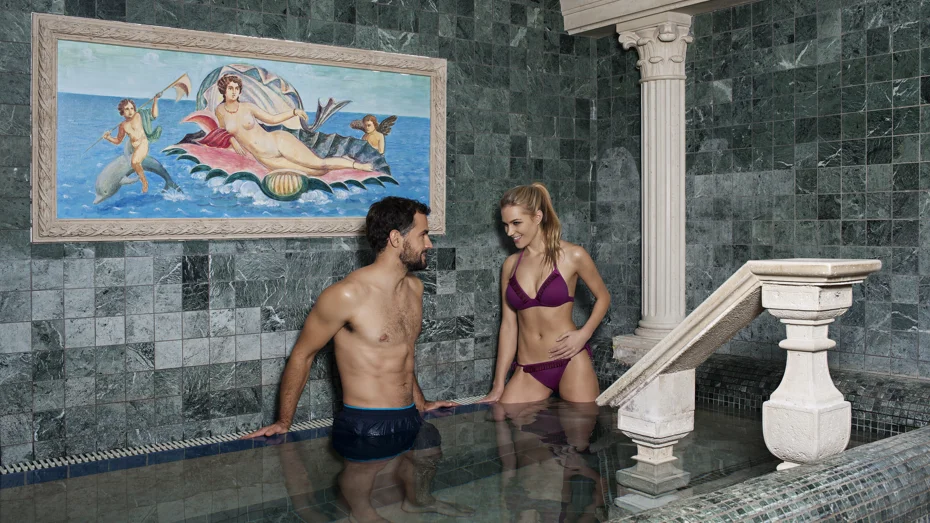 Moški in ženska se v kopalkah pogovarjata ob robu notranjega termalnega bazena
