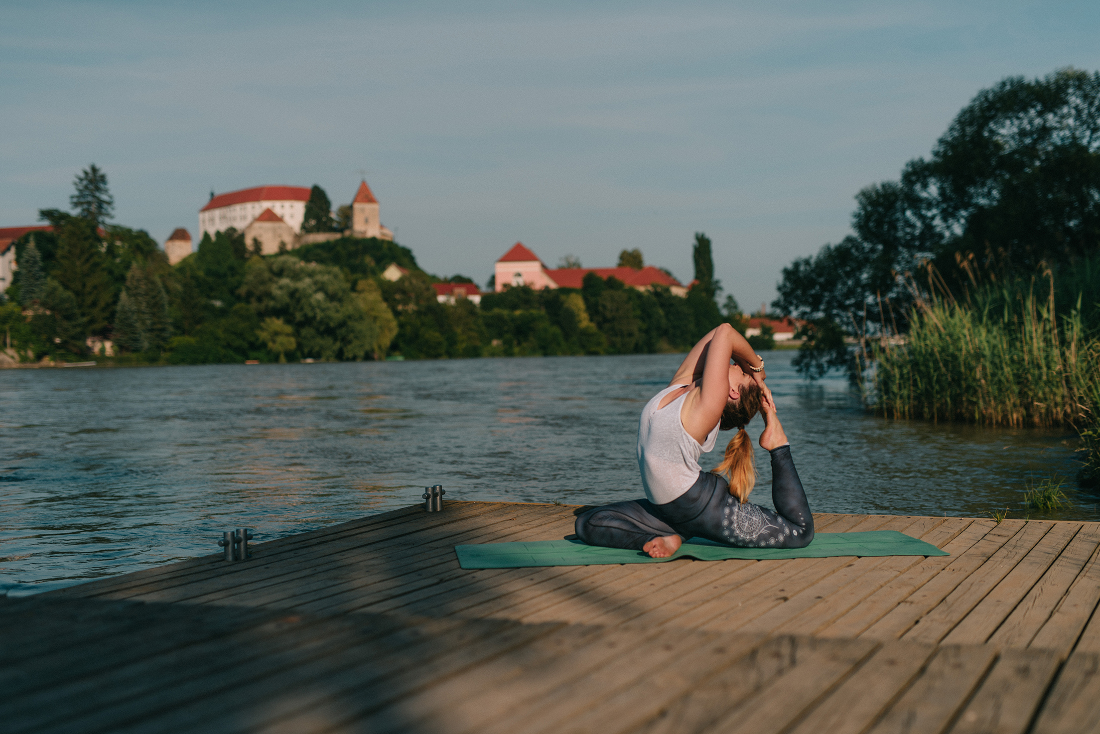 Ženska v športnih oblačilih na zeleni podlogi izvaja jogo ob bližnjem jezeru