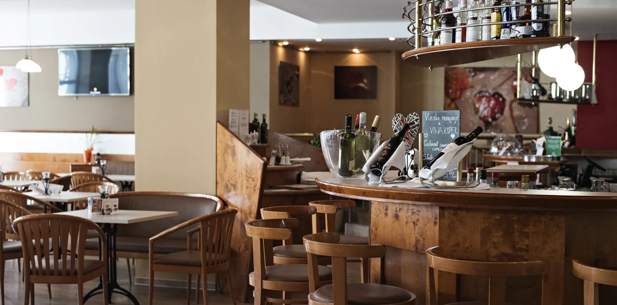 Prijeten hotelski bar z lesenim pultom, visokimi stoli, mizami in televizijo v ozadju