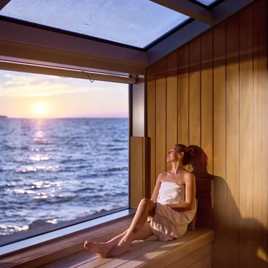 Ženska v brisači leži na leseni konstrukciji notranje termalne riviere in si čez okno ogleduje morje