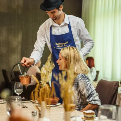 Ženska sedi za hotelsko mizo, natakar z modrim predpasnikom pa ji razlaga prekmursko jed