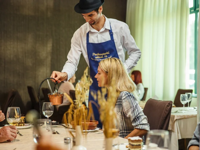 Ženska sedi za hotelsko mizo, natakar z modrim predpasnikom pa ji razlaga prekmursko jed