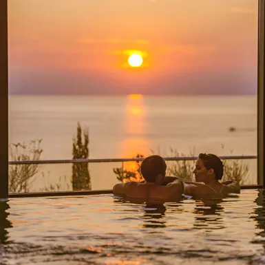 Moški in ženska se naslanjata ob rob notranjega bazena in se pogovarjata ob pogledu na sončni zahod