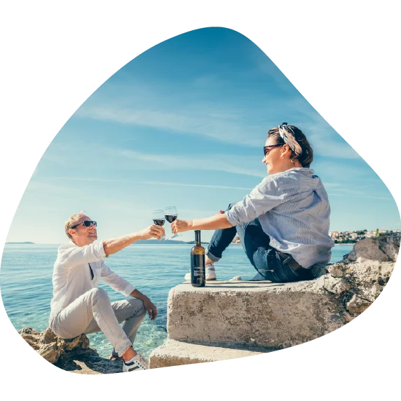 Moški in ženska srednjih let sedita na kamnih ob morju in nazdravljata z vinom