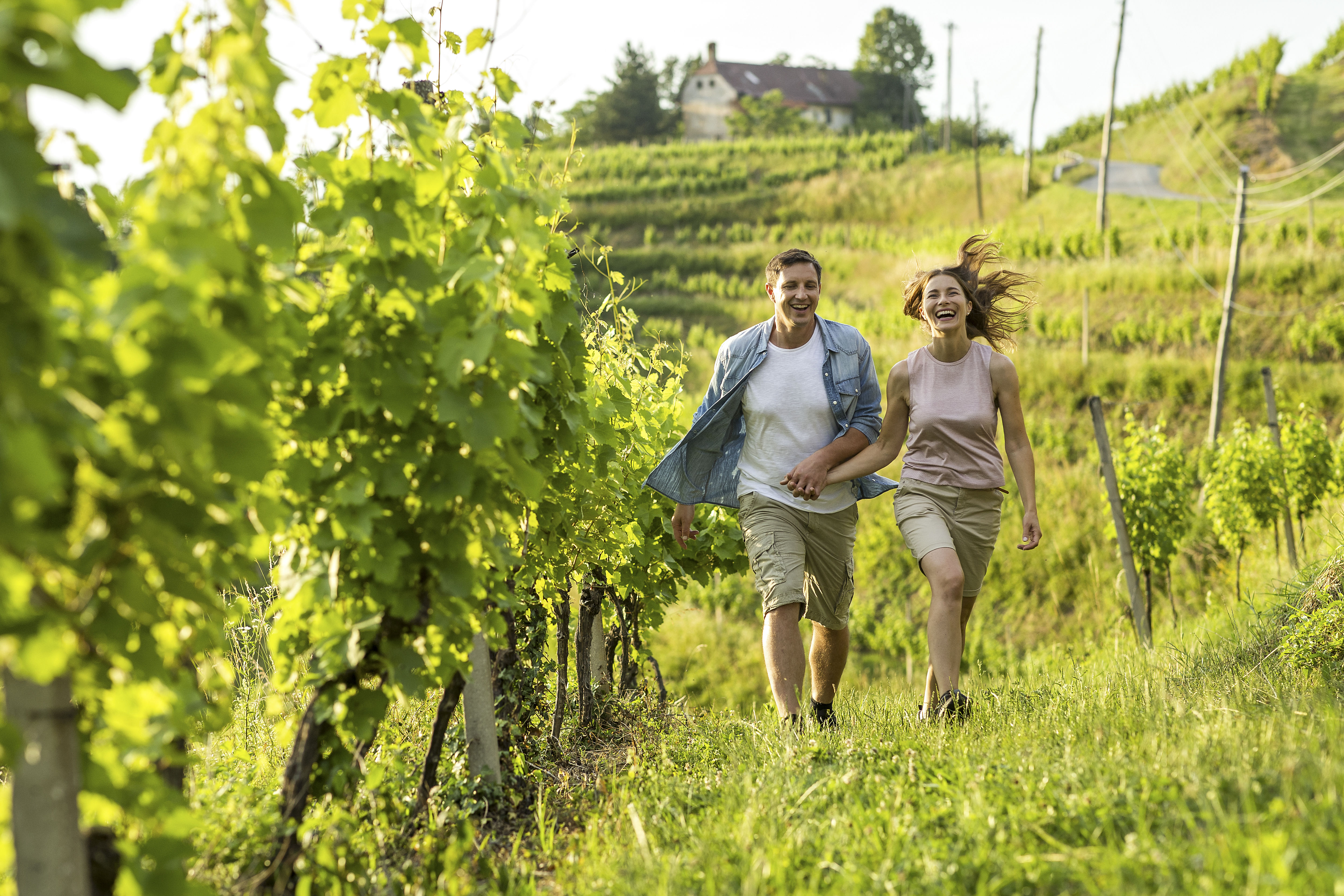 Moški in ženska v športnih oblačilih se nasmejano držita za roke in sprehajata med zelenimi vinogradi