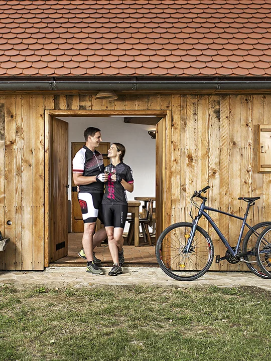 Mlad kolesarski par, oblečen v kolesarska oblačila, stoji pred leseno hišico in se pogovarja