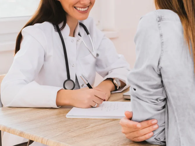 Nasmejana zdravnica v beli halji sprašuje pacientko in si zapisuje ugotovitve