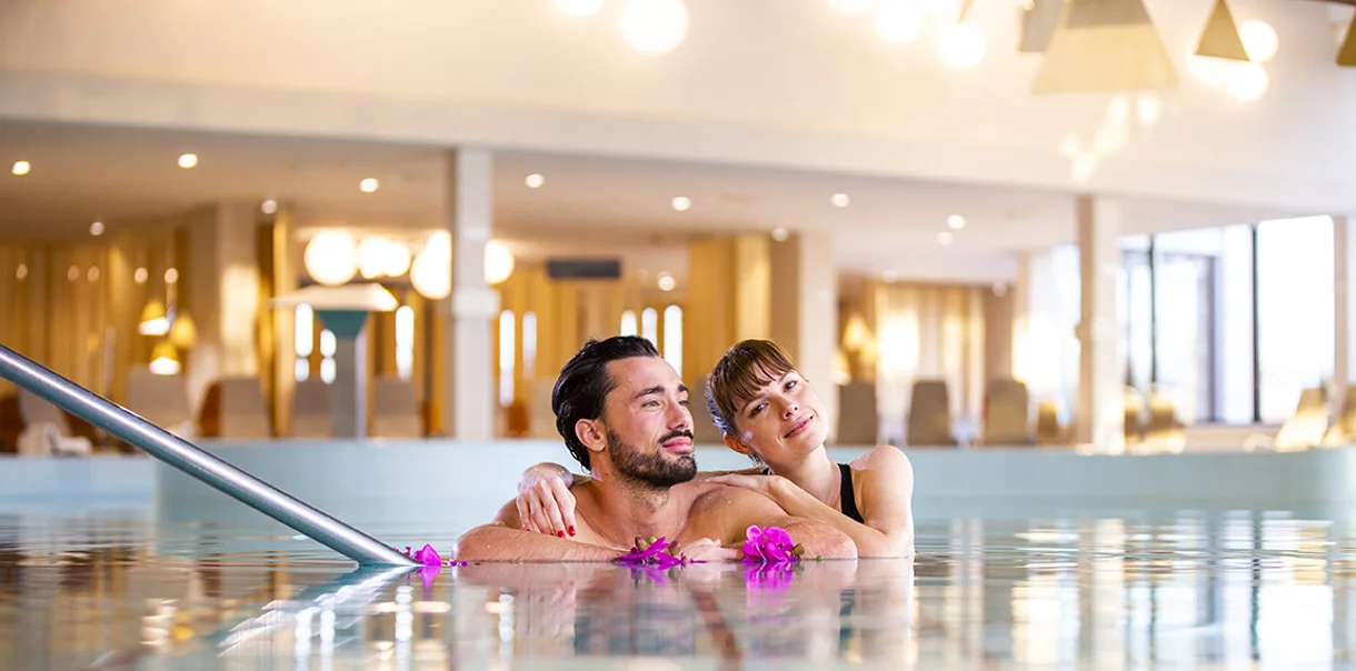 Moški in ženska se objemata in uživata v notranjem termalnem bazenu z roza rožami v vodi