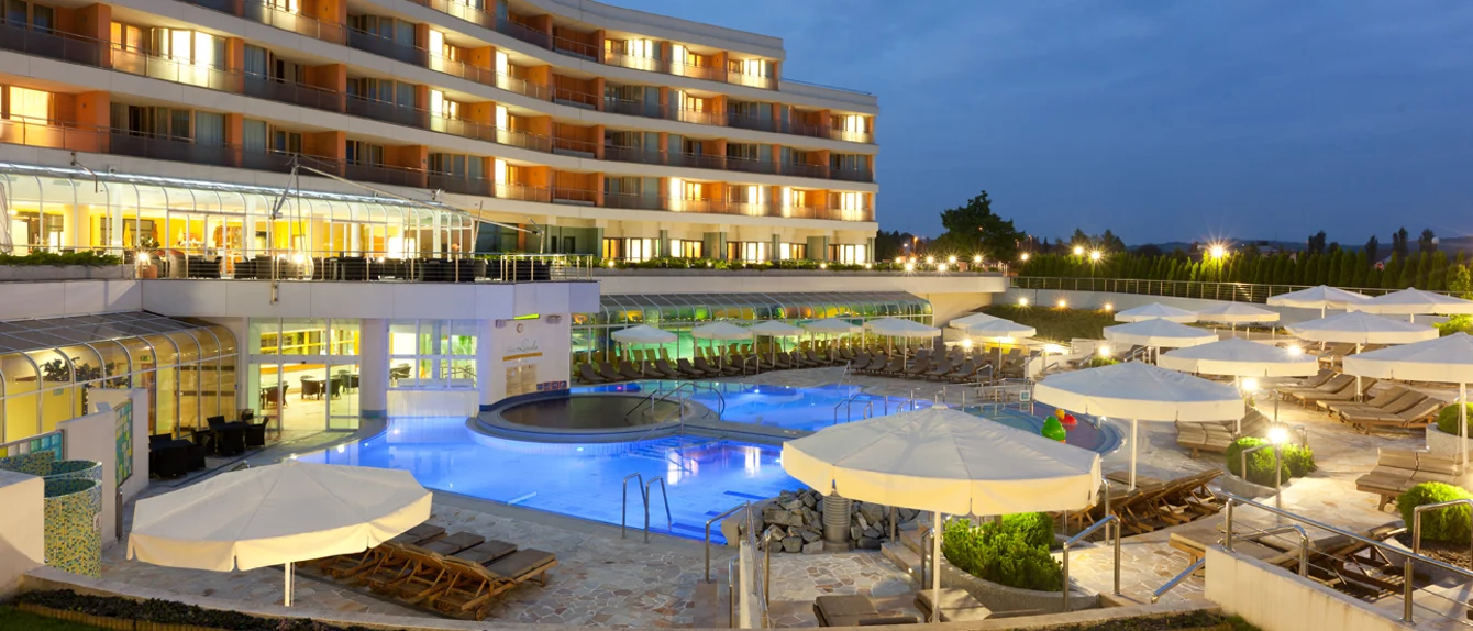 Pogled na osvetljen zunanji bazen, bele dežnike, ležalnike, teraso in hotel