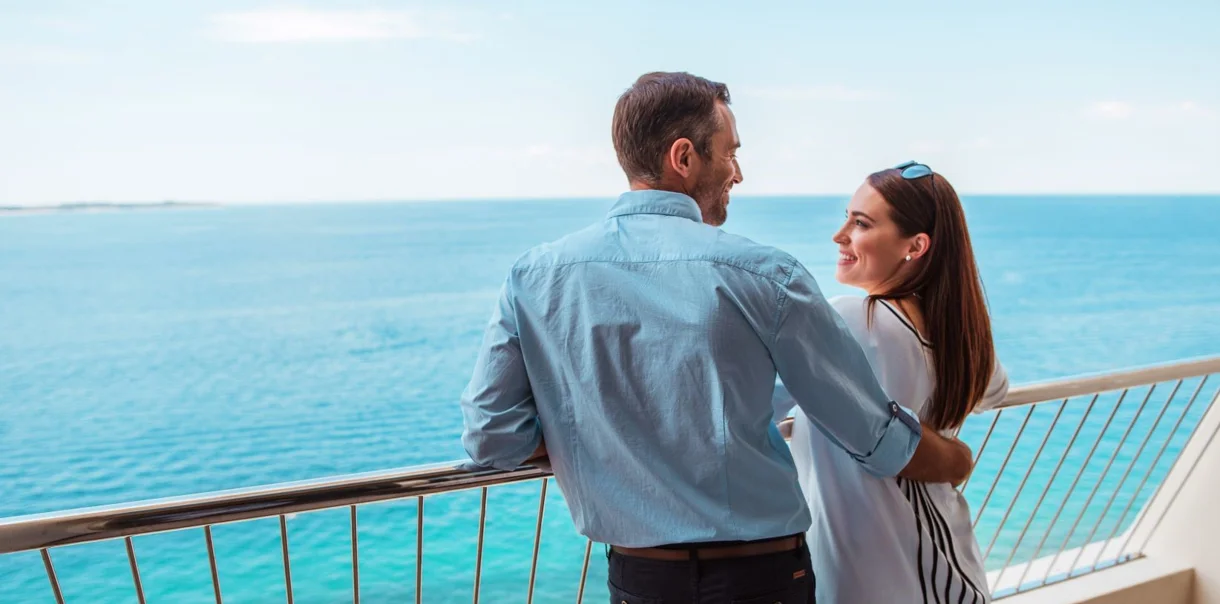 Moški in ženska se nasmejano objemata ob ograji terase s pogledom na morje