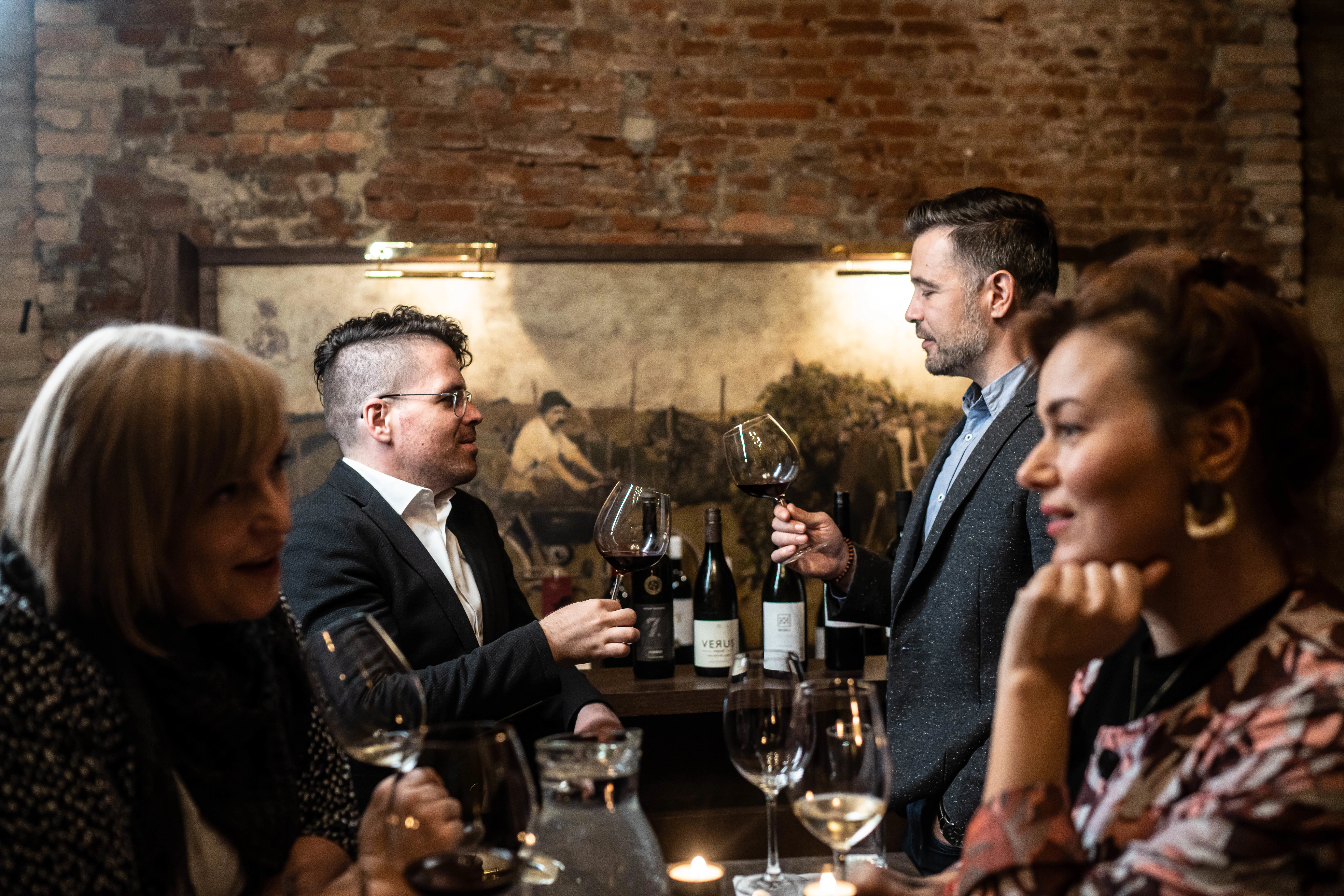 Dva moška in dve ženski se v vinski kleti pogovarjajo za dvema mizama s kozarcem vina v roki